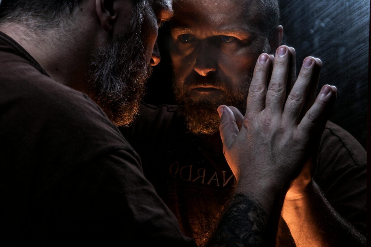 A bearded man with a stern expression leans against a mirror in low-light setting.