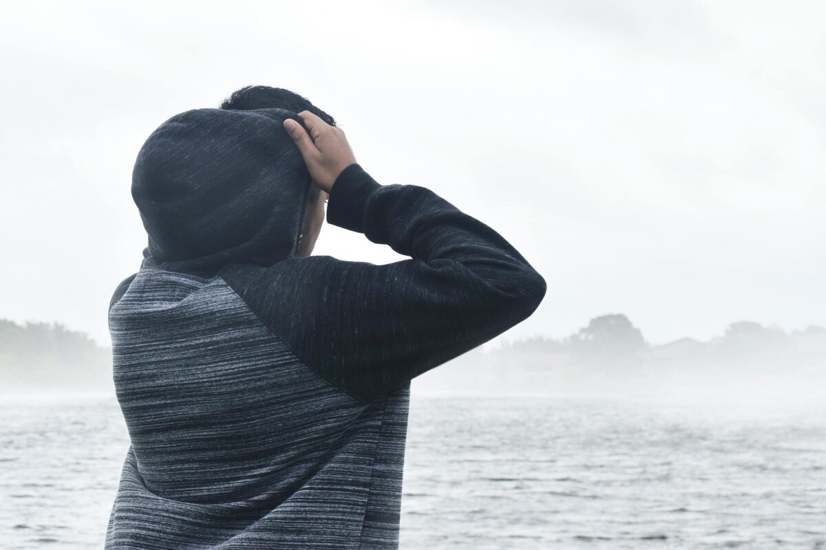A person wearing a hoodie stands by a misty lake, gazing at the foggy horizon.
