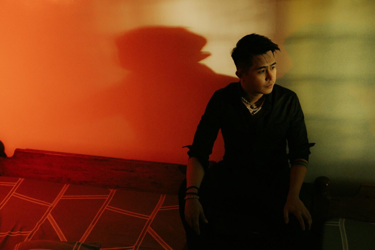 A reflective young man sits in a dimly lit room, casting a shadow against a warm-toned wall.
