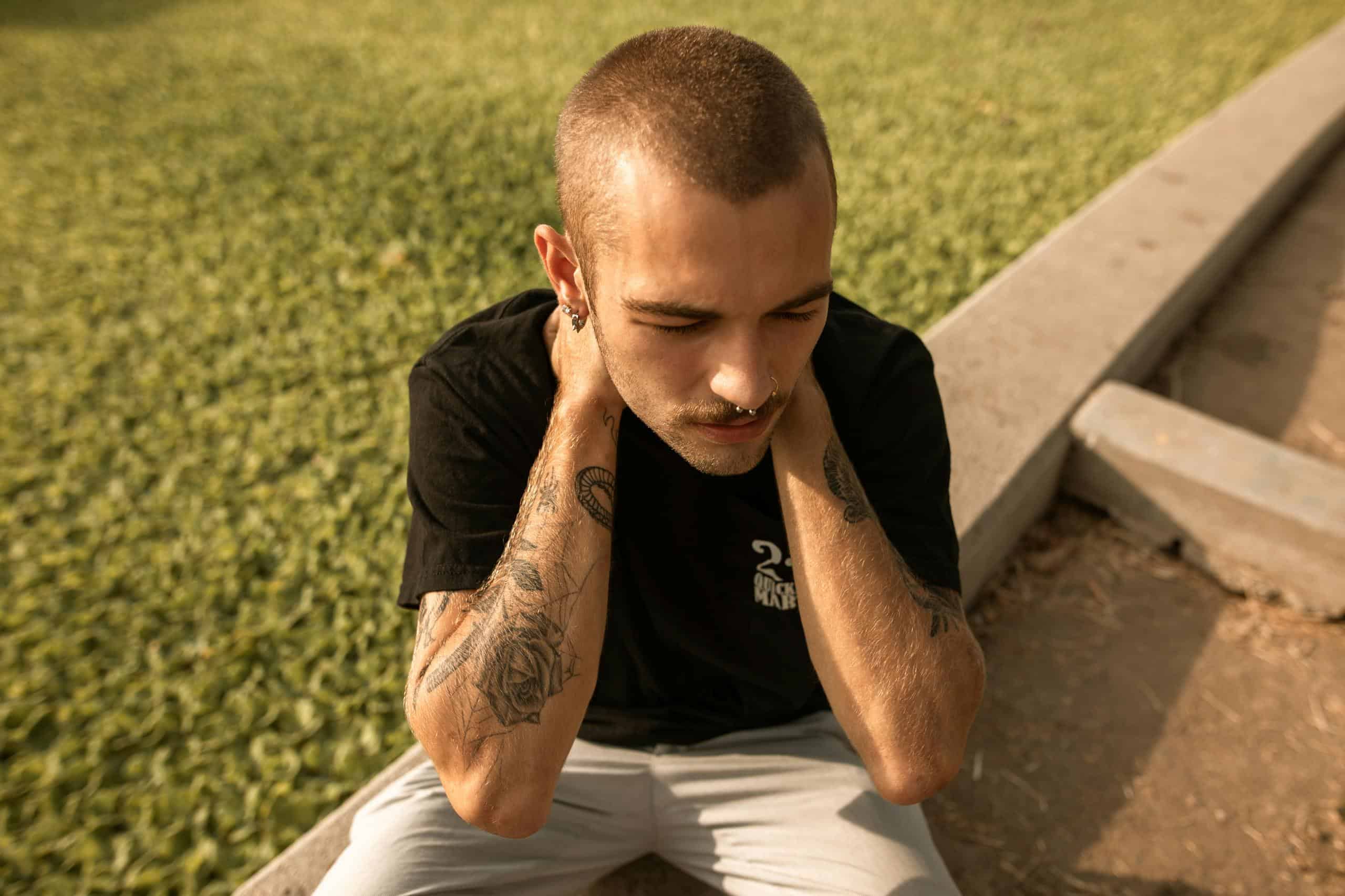A young tattooed man sits outdoors, deep in thought, hands on neck.