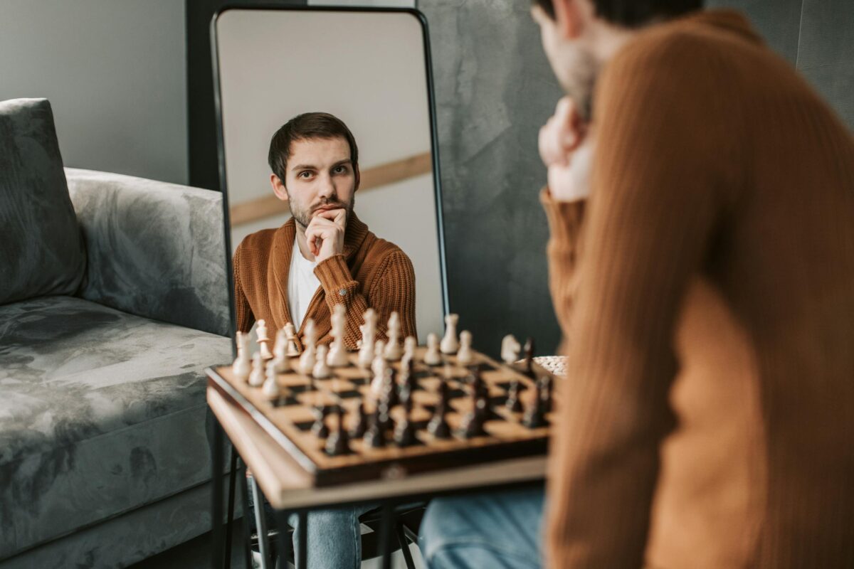 Man in brown sweater engages in chess, captured in thoughtful reflection through a mirror indoors.