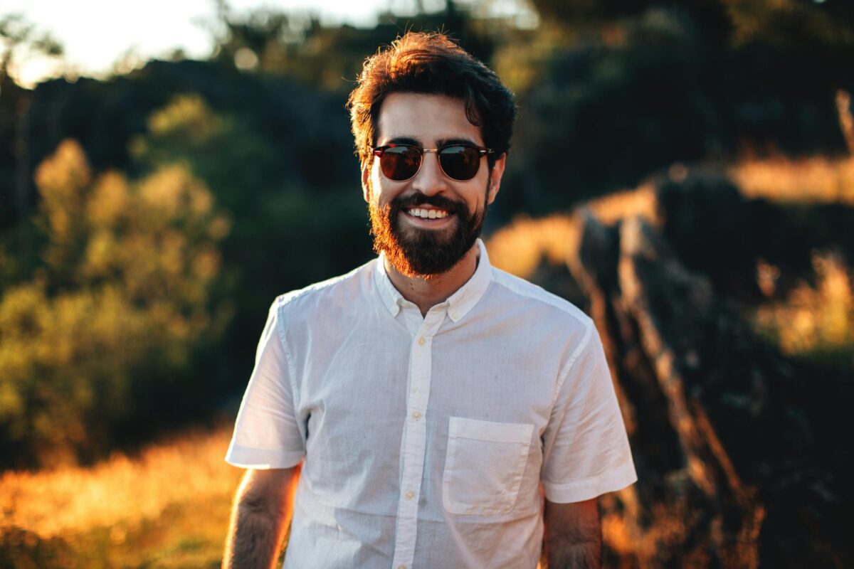 Portrait of a bearded man in sunglasses smiling outdoors with sunlight and natural background.