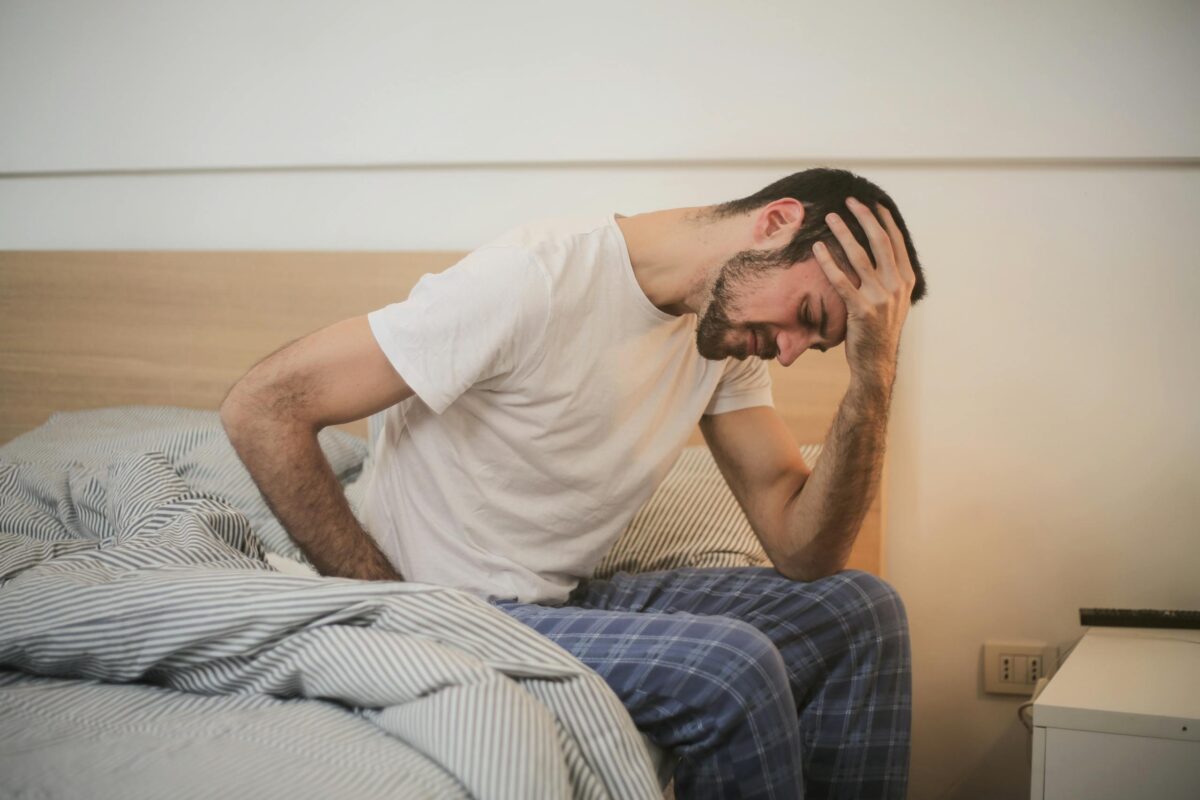 Side view of male in sleepwear sitting on bed with closed eyes after awakening and suffering from migraine while touching head and grimacing in pain
