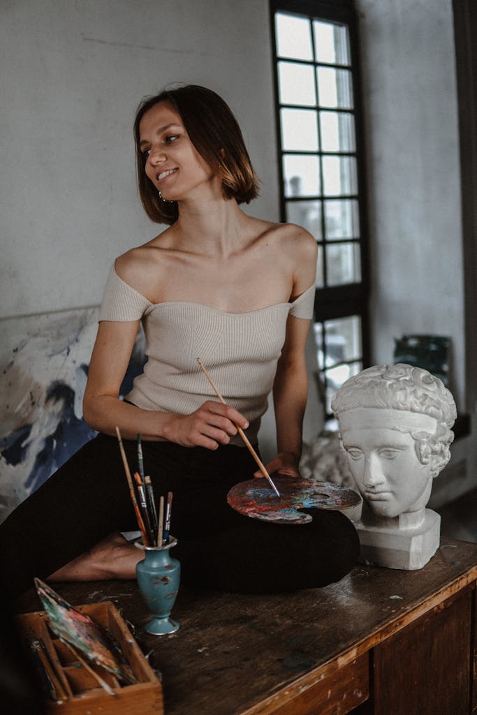 Artist sitting indoors with paintbrushes and a sculpture, creating art.
