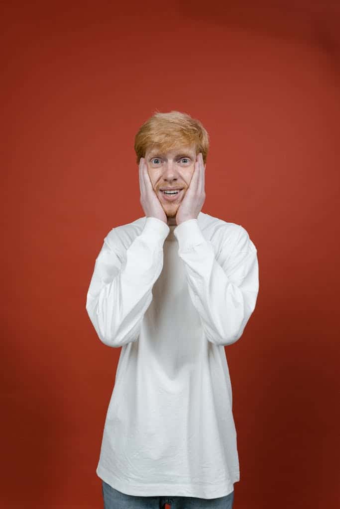 Portrait of a surprised man with blonde hair in a white sweater posing against a red studio background.