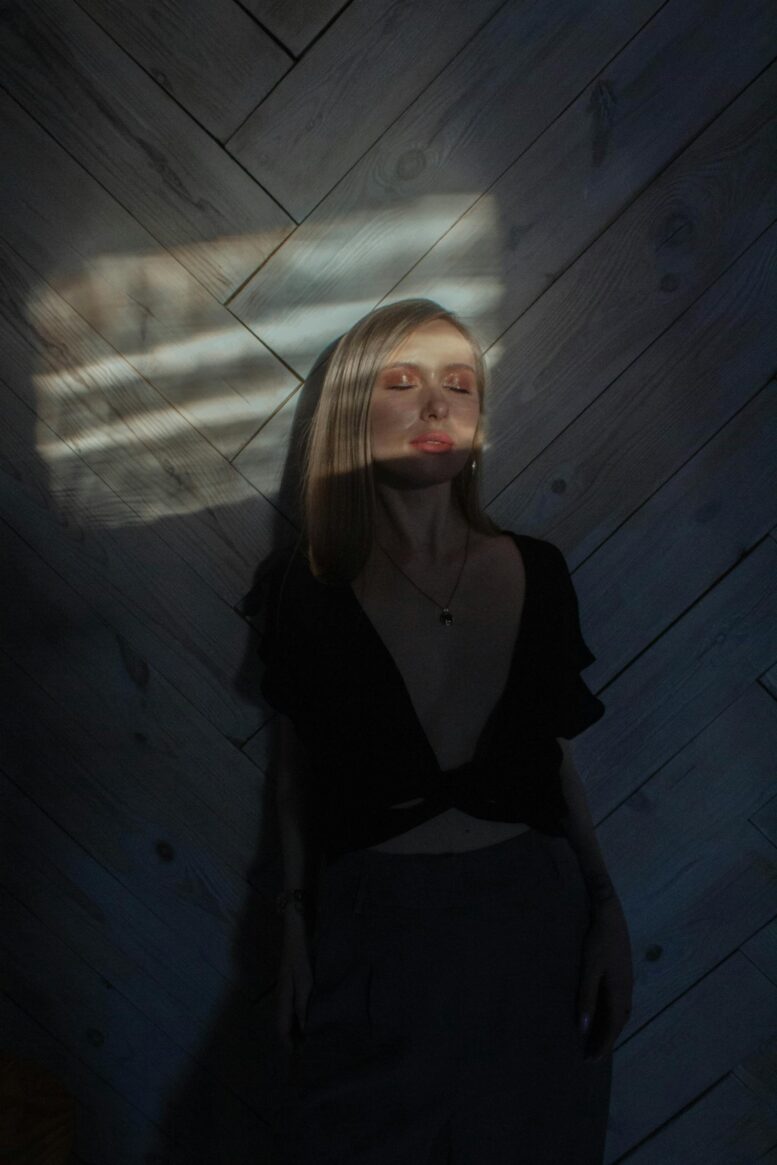 Young mindful female in trendy apparel standing with closed eyes against wall with shadows in sun rays